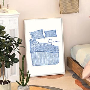 May include: A framed art print featuring a blue line drawing of a bed with pillows and bedding. The text "Let's Stay In Bed" is written in blue script. The artwork is displayed on a white wall.