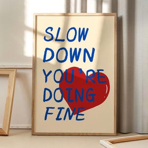 May include: A framed print with a cream background and blue text that reads "Slow Down You're Doing Fine". A red heart is behind the text.