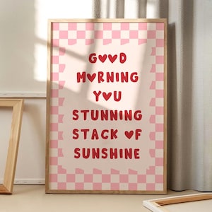 Puede incluir: Una impresión artística enmarcada con un fondo rosa claro y un borde a cuadros rosa y blanco. El texto rojo dice "GOOD MORNING YOU STUNNING STACK OF SUNSHINE" con símbolos de corazón. El marco es de color marrón claro.