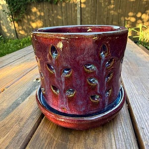 May include: A cylindrical ceramic planter with a deep burgundy and purple glaze featuring a glossy finish. The exterior is decorated with numerous small, leaf-shaped cutouts arranged in vertical rows. It includes a matching circular drip tray.