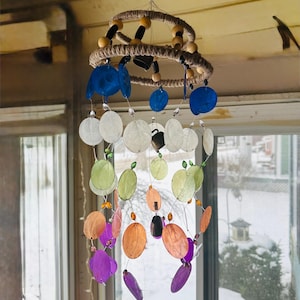 Puede incluir: Un carillón de viento decorativo con un diseño en espiral. Presenta piezas en forma de concha de color azul, blanco, verde, naranja y morado, suspendidas de un anillo envuelto en cuerda. También incorpora cuentas de madera y pequeñas campanas.