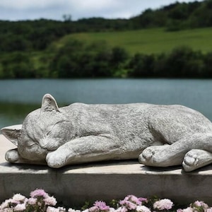 May include: A gray concrete cat statue sleeping on a stone ledge with a lake and green hills in the background.