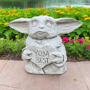 Pu&ograve; includere: Una statua di pietra grigia di un personaggio della saga di Star Wars, che tiene un cartello a forma di cuore che recita "Yoda Best". La statua &egrave; circondata da piante verdi e fiori.