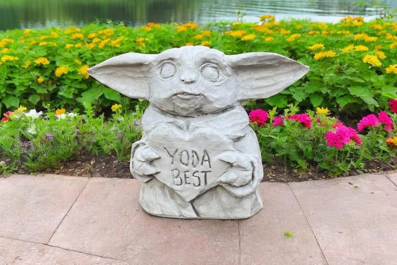 Pu&ograve; includere: Una statua di pietra grigia di un personaggio della saga di Star Wars, che tiene un cartello a forma di cuore che recita "Yoda Best". La statua &egrave; circondata da piante verdi e fiori.