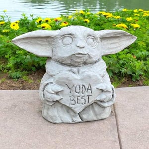 Pu&ograve; includere: Una statua di pietra grigia di un personaggio del franchise di Star Wars, che tiene un cartello a forma di cuore che dice "Yoda Best".