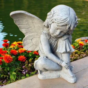 May include: A gray concrete fairy statue with large wings, seated with its head resting on its knees. The fairy is wearing a dress and has curly hair. The statue is placed near a pond with colorful flowers in the foreground.