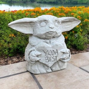 Pu&ograve; includere: Una statua di cemento grigio di un personaggio di Baby Yoda che tiene un cartello a forma di cuore che dice "Yoda Best". La statua &egrave; seduta su un patio di cemento grigio.