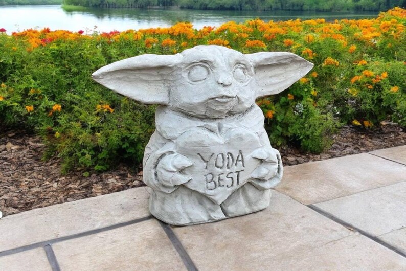 Pu&ograve; includere: Una statua di cemento grigio di un personaggio di Baby Yoda che tiene un cartello a forma di cuore che dice "Yoda Best". La statua &egrave; seduta su un patio di cemento grigio.