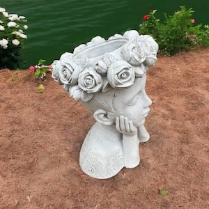 May include: A white concrete planter shaped like a girl's head with a flower crown. The planter is resting on a bed of brown mulch.