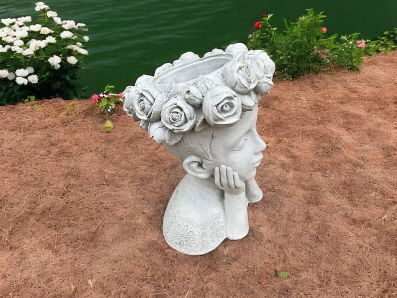 May include: A white concrete planter shaped like a girl's head with a flower crown. The planter is resting on a bed of brown mulch.