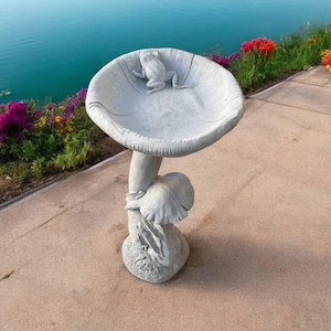 May include: A gray concrete bird bath shaped like a mushroom with a frog on top. The bird bath is sitting on a concrete patio.