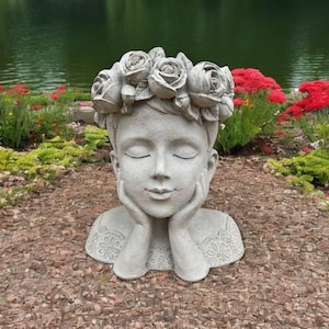 May include: A gray concrete sculpture of a person's head with a flower crown. The person has their eyes closed and their hands are resting on their chin. The sculpture is sitting on a bed of brown mulch.
