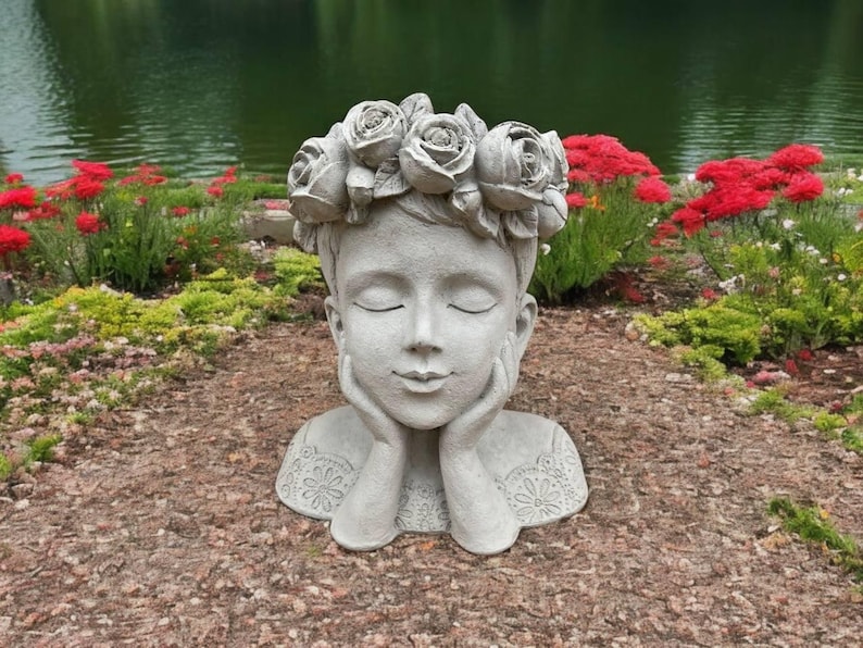 May include: A gray concrete sculpture of a person's head with a flower crown. The person has their eyes closed and their hands are resting on their chin. The sculpture is sitting on a bed of brown mulch.