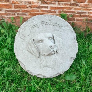 May include: A round, gray stone garden stepping stone with the words "My Pointer" and a heart carved into the surface. The stone features a detailed relief of a dog's head, set against a backdrop of green grass and a brick wall.