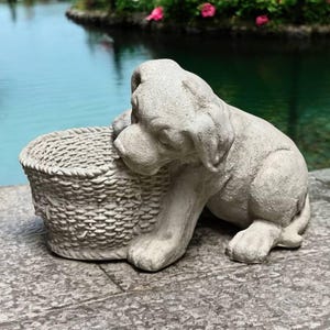 May include: A gray concrete statue of a dog sitting next to a woven basket. The statue is outdoors on a stone surface near a pond.