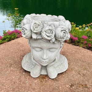 May include: A white concrete planter shaped like a woman's head with a crown of roses around the top. The planter is sitting on a gray rock surface.