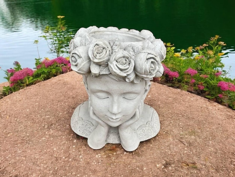May include: A white concrete planter shaped like a woman's head with a crown of roses around the top. The planter is sitting on a gray rock surface.