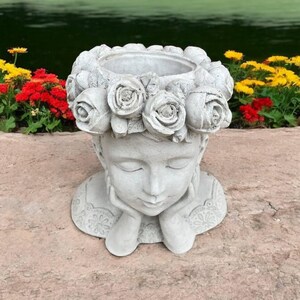 May include: A white concrete planter shaped like a woman's head with a wreath of roses around the top. The woman's eyes are closed and she is holding her chin with both hands.