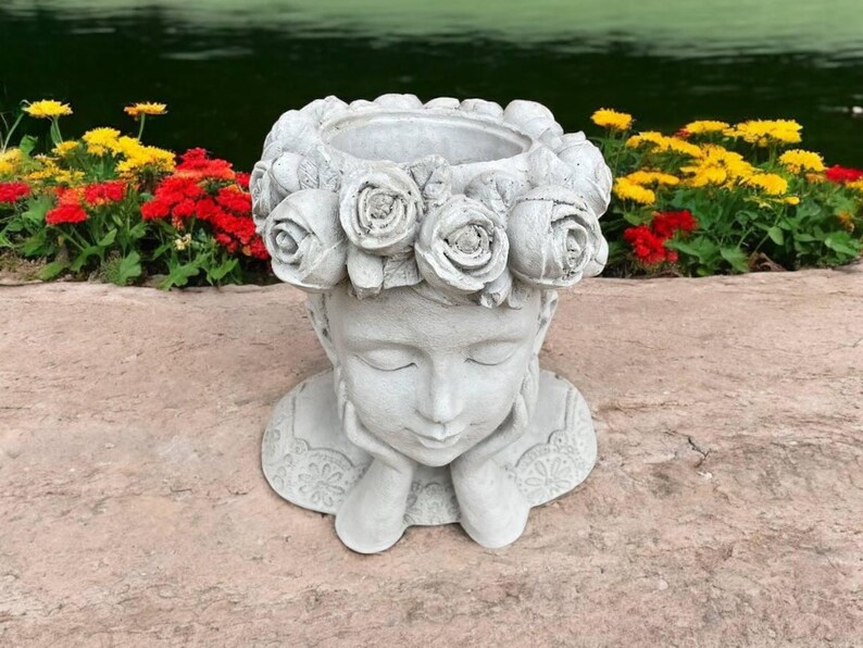 May include: A white concrete planter shaped like a woman's head with a wreath of roses around the top. The woman's eyes are closed and she is holding her chin with both hands.