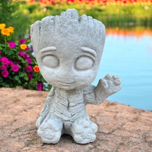 May include: A gray concrete planter shaped like a character with large eyes and a friendly expression. The planter has a textured surface and is designed to hold small plants or flowers. The character is waving with one hand.