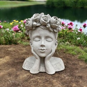 May include: A gray concrete planter shaped like a woman's head with a flower crown. The woman has her eyes closed and her hands are resting on her chin. The planter is sitting on a bed of brown dirt.