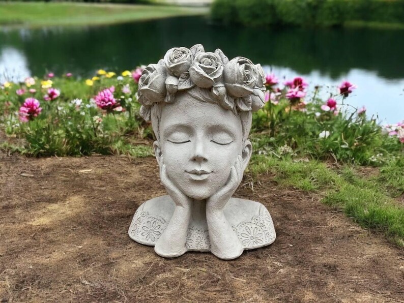 May include: A gray concrete planter shaped like a woman's head with a flower crown. The woman has her eyes closed and her hands are resting on her chin. The planter is sitting on a bed of brown dirt.
