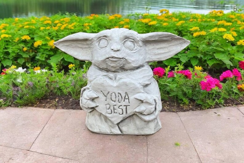 Pu&ograve; includere: Una statua di pietra grigia di un personaggio della saga di Star Wars, che tiene un cartello a forma di cuore che recita "Yoda Best". La statua &egrave; circondata da piante verdi e fiori.