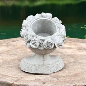 May include: A gray concrete planter shaped like a woman's bust with a floral design around the top. The planter has a decorative lace pattern around the base.