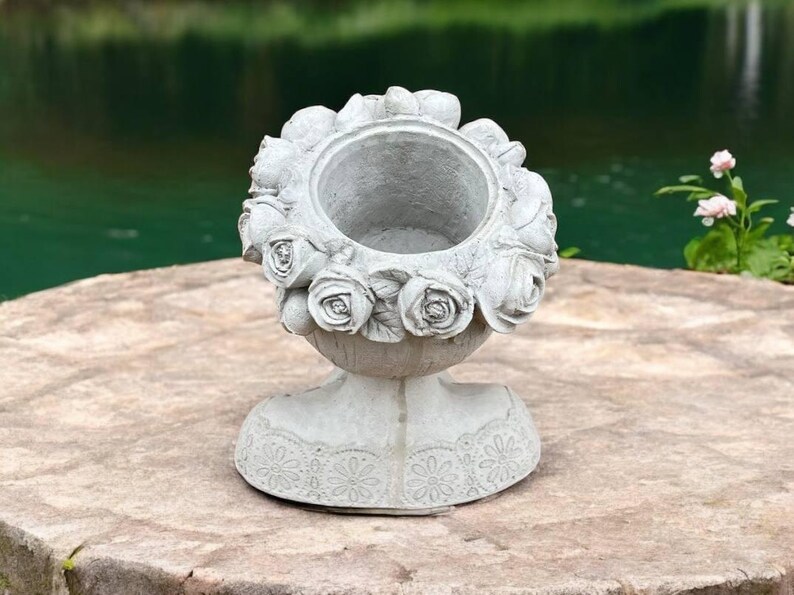 May include: A gray concrete planter shaped like a woman's bust with a floral design around the top. The planter has a decorative lace pattern around the base.