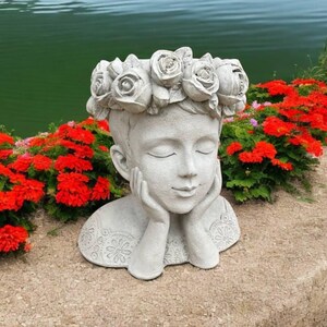 May include: A white concrete sculpture of a person's head with a flower crown. The sculpture is resting on a bed of red flowers.