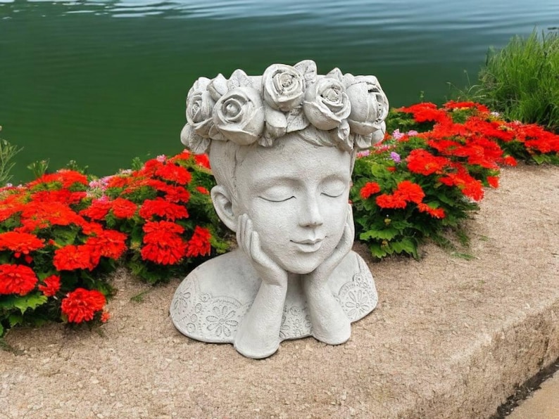 May include: A white concrete sculpture of a person's head with a flower crown. The sculpture is resting on a bed of red flowers.
