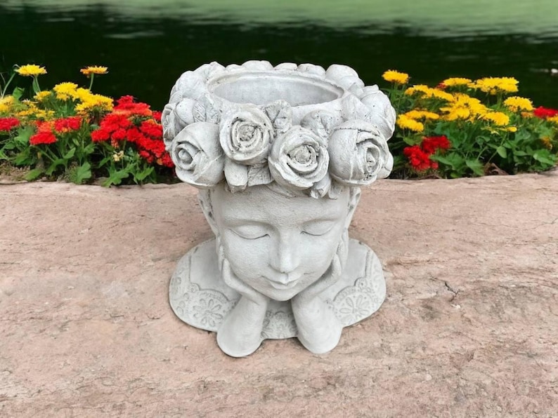 May include: A white concrete planter shaped like a woman's head with a wreath of roses around the top. The woman's eyes are closed and she is holding her chin with both hands.