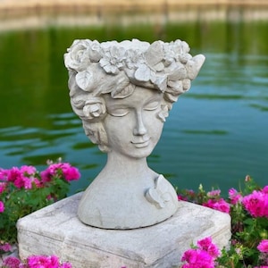 May include: A gray concrete planter shaped like a woman's head with a floral crown. The planter is sitting on a stone pedestal in front of a pond with pink flowers.