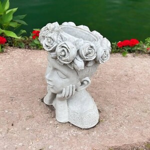 May include: A gray concrete planter shaped like a woman's head with a flower crown. The planter is designed to hold plants or flowers.