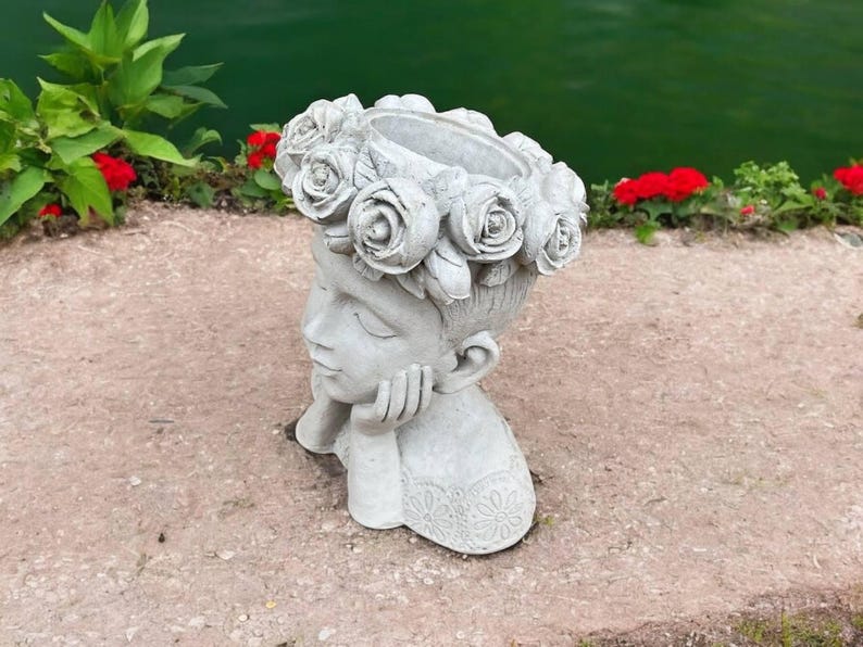 May include: A gray concrete planter shaped like a woman's head with a flower crown. The planter is designed to hold plants or flowers.