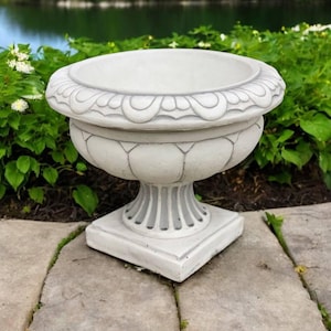 May include: A white ceramic planter with a decorative rim and a pedestal base. The planter is sitting on a stone patio.