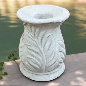 May include: A light gray, decorative outdoor planter with a wide, circular opening. The planter features a detailed leaf design carved into the sides. The base is round, and the planter is approximately 12 inches tall.