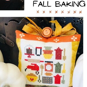 May include: A small, square, white cushion with a cross-stitch design of various autumn baking icons. The cushion is decorated with a yellow ribbon and a wooden button. The cushion is surrounded by white pumpkins.