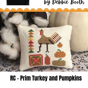 Puede incluir: Una pequeña almohada blanca con un diseño de punto de cruz de un pavo y calabazas. La almohada está sobre un fondo de bolas de algodón blancas y una tela estampada marrón y blanca. El texto "RC - Prim Turkey and Pumpkins" está en la parte inferior de la imagen.