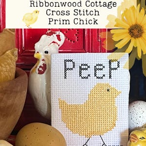 Peut inclure: Motif de point de croix représentant un poussin jaune avec le mot "peep" au-dessus. Le motif est encadré et exposé sur une étagère avec d'autres décorations de Pâques, notamment un coq en céramique, un œuf brun et une figurine de poussin jaune.