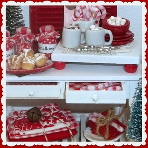 May include: A white table with two drawers, a red and white checkered tablecloth, and a white ceramic countertop. The countertop has a red and white candy cane, a red and white jar labeled "mallows", and two white mugs with red and white candy canes. The table is decorated with red and white Christmas decorations.