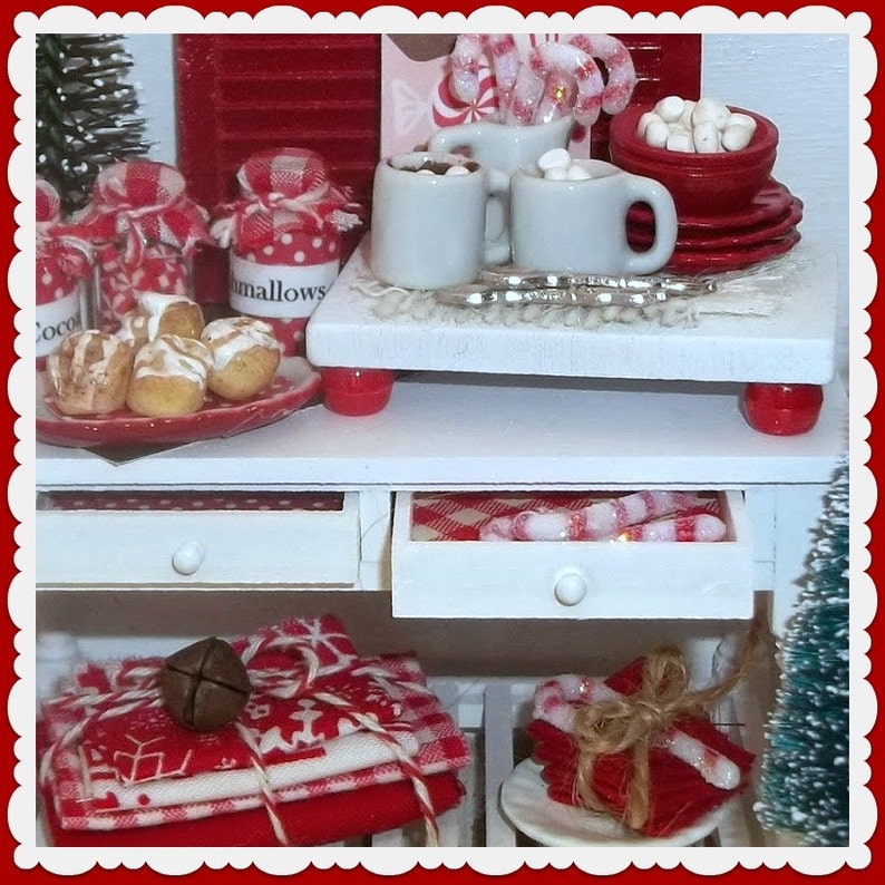 May include: A white table with two drawers, a red and white checkered tablecloth, and a white ceramic countertop. The countertop has a red and white candy cane, a red and white jar labeled "mallows", and two white mugs with red and white candy canes. The table is decorated with red and white Christmas decorations.
