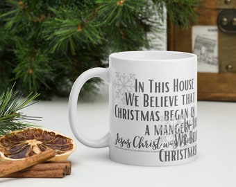 May include: White ceramic mug with a black and gray design that reads "In this house we believe that Christmas began in a manger..." The mug has a snowflake design on the side.