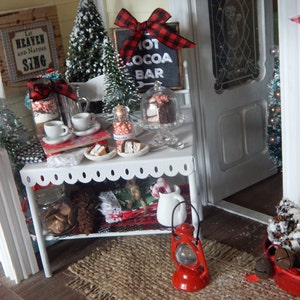 May include: A miniature dollhouse scene with a white, scalloped-edge table with a red and black checkered ribbon, a red lantern, and a basket of pine cones. The table is decorated with miniature Christmas trees, a sign that says "Hot Cocoa Bar", and a variety of miniature desserts.