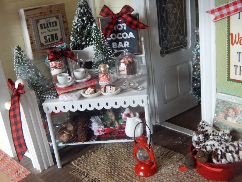 May include: A miniature dollhouse scene with a white, scalloped-edge table with a red and black checkered ribbon, a red lantern, and a basket of pine cones. The table is decorated with miniature Christmas trees, a sign that says "Hot Cocoa Bar", and a variety of miniature desserts.