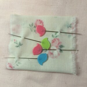 Op de afbeelding: Vier kleurrijke plastic naalden met metalen naalden zijn vastgezet in een witte stof met een bloemenpatroon. De naalden zijn bladvormig en zijn roze, groen, blauw en lichtroze.