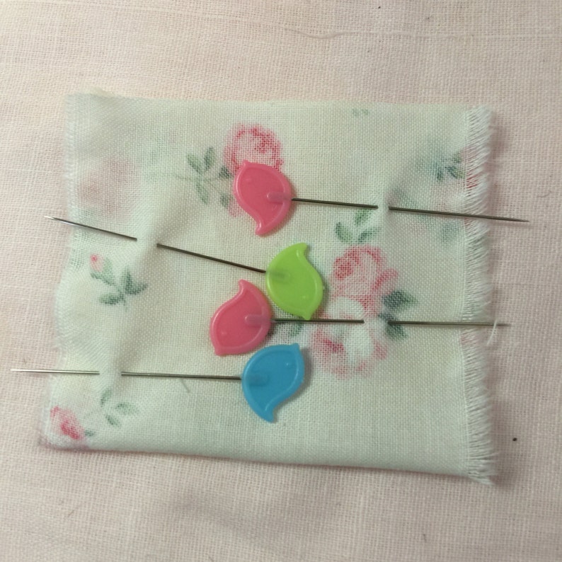 Op de afbeelding: Vier kleurrijke plastic naalden met metalen naalden zijn vastgezet in een witte stof met een bloemenpatroon. De naalden zijn bladvormig en zijn roze, groen, blauw en lichtroze.
