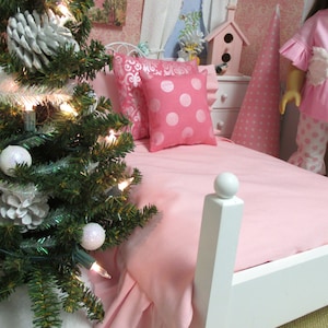 May include: A pink doll bed with a white headboard and footboard. The bed is made with a pink sheet and a pink and white polka dot pillow. There is a white and pink patterned pillow on the bed as well. A small Christmas tree with white ornaments and lights is in the foreground.