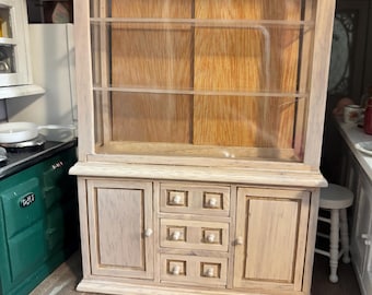 May include: A miniature wooden hutch with a glass-fronted upper cabinet and a lower cabinet with doors and drawers. The hutch is unfinished, showing the natural wood grain. The piece is part of a dollhouse kitchen setting.