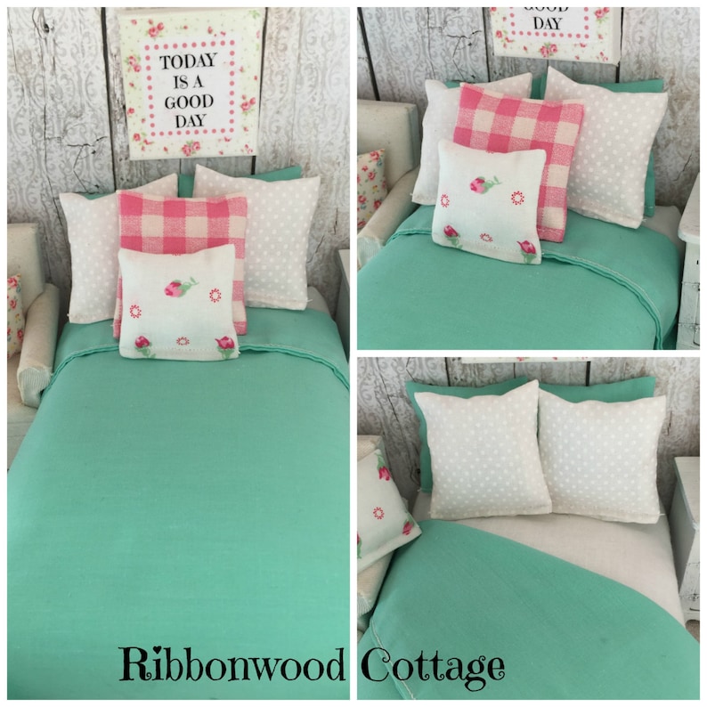 May include: A doll-sized bed with a teal green duvet and white and pink patterned pillows. The pillows have embroidered flowers. The sign above the bed reads "Today is a good day".